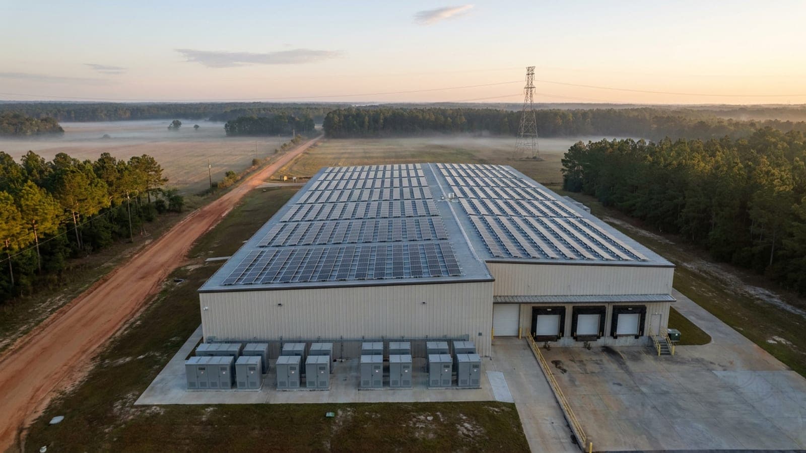 Aerial view of a rooftop solar array and battery storage cabinets on a commercial building in coastal Georgia at sunrise, with transmission lines beyond